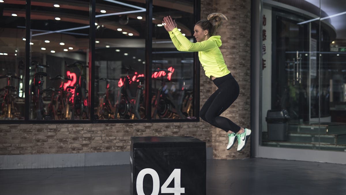 Fit young woman in sports wear executing a box jump indoors, promoting fitness and health in a modern urban gym.